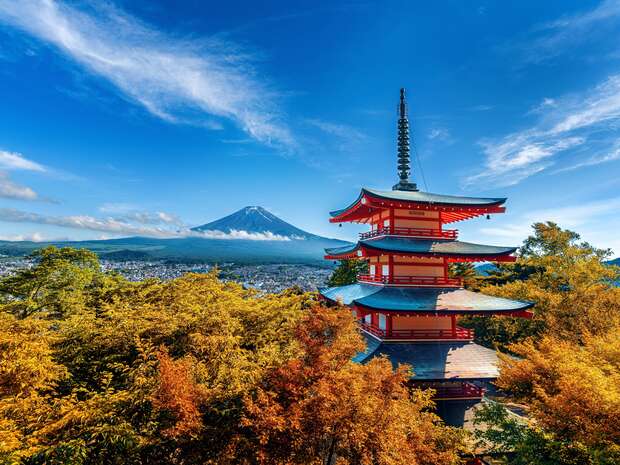 A view of Chureito Pagoda in the foreground with Mount Fuji in the distance. Autumn foliage surround the building, and a blue sky is overhead.